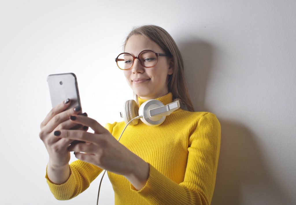 photo of smiling woman in yellow turtleneck sweater with headphones on her neck using her phone
